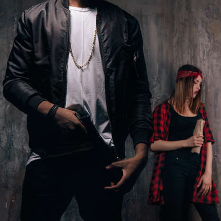 Guy with gun and girl with alcohol. Criminal teenagers on dark background. Gang members lifestyleの写真素材