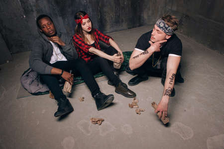 Group of homeless gung members gets drunk. Youth addiction problem. Tired drunk caucasian girl holding alcohol bottle, sad and depressed guys with tattoo sits near . Social problem conceptの写真素材