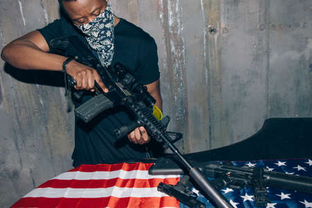 Black sniper aims at the rifle. Mercenary man with weapon in hand on dark gray background. Outlaw,...