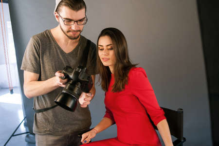 Photographer communicate with model on photoshoot. Man with camera show pictures to woman in red...
