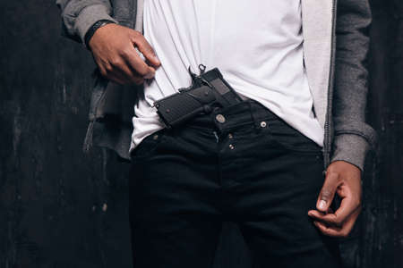 Unrecognizable black man threatens with a gun closeup studio shoot. Ghetto gangster with weapon on dark background. Outlaw, ghetto, murderer, robbery conceptの写真素材