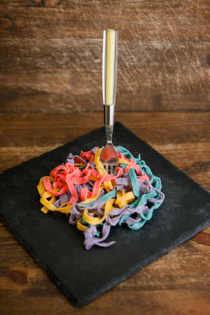 Colorful tagliatelle with fork on black plate, wooden background closeup, culinary conceptの写真素材