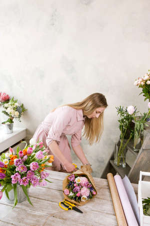 Decorator pack festive bouquet in wrapping paper. Young beautiful florist make floristry assemble with pink peony and wildflowers in workshop on wooden background. Woman at workの写真素材
