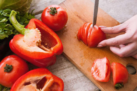 Tomatoes cutting on wooden desk by unrecognizable woman. Red pepper, and parsley nearby on table, cooking process of fresh vegetables saladの写真素材