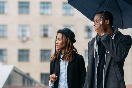 Thoughtful fashion pedestrians. Rainy mood. Young black couple with umbrella , happy African American, perspective conceptの写真素材