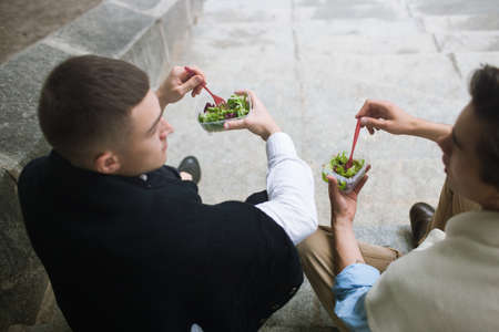 Healthy lunch break outdoors. Nutrition diet. Unrecognizable young men, two male friends in selective focus, modern youth, food conceptの写真素材