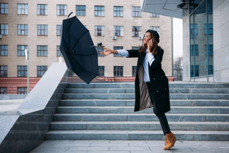 Stormy weather. Black girl catch flying umbrella while speaking on mobile phone on street, airiness conceptの写真素材