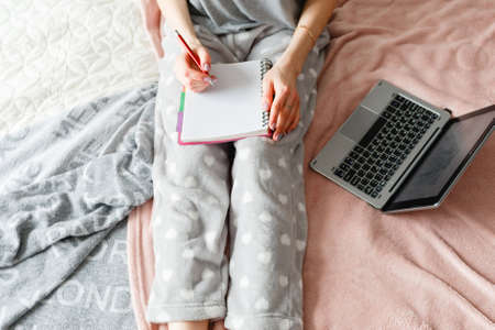 Unrecognizable woman makes notes in writing book, laptop nearby. Education, memories and thoughts, planning and to-do list for day, close up top viewの写真素材