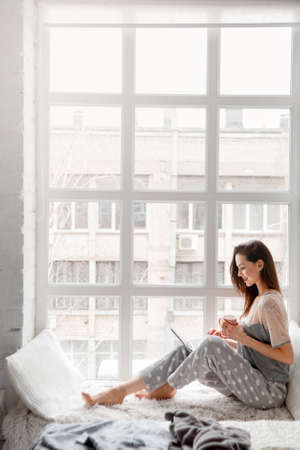 Smiling woman sitting near white window and chatting on laptop. Working start from the morning, making notes, social life, freelance worker concept. Side view picture with free spaceの写真素材