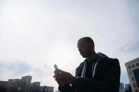Social media communication outdoors. Black male typing with smartphone. Sky background with free space, modern technology, street styleの写真素材