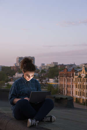 Freelance work outdoors. Self-education for girl. Happy young female with laptop on roof in focus on foreground, modern social media communication, urban backgroundの写真素材