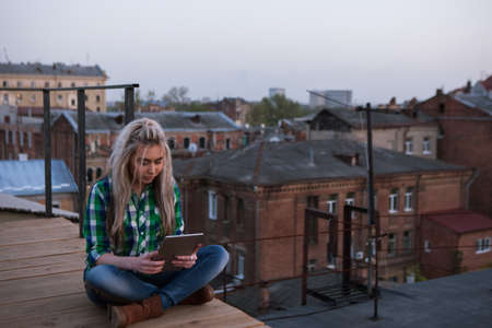 Freelance work outdoors. Modern girl on roof. Social media communication, gadget addiction , stylish female with tablet in focus on foreground, urban backgroundの写真素材