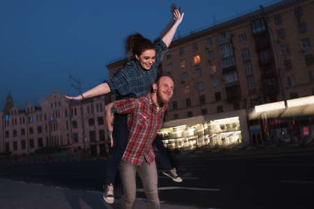 Imaginary flight. Happy love couple in motion. Blurred urban background, cheerful man giving girl piggyback ride, flying smiliey female, fun conceptの写真素材