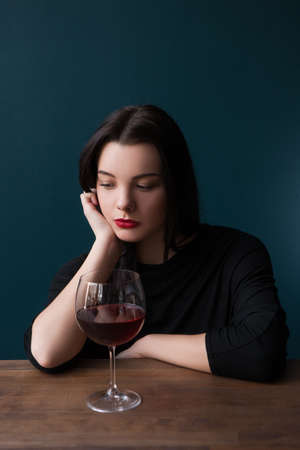 Lonely female in bar. Melancholic mood. Sad woman with red wine on blue background with free space,...