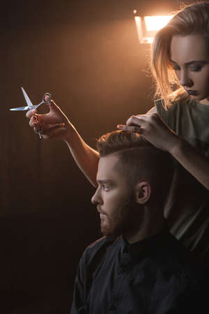 Female hairdresser cutting modern hipster hair. Hairdressing salon in dark place, stylish young male...