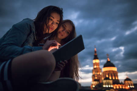 Friends with a tablet in a night city. Girls communicate in a social network, make purchases online, work remotelyの写真素材