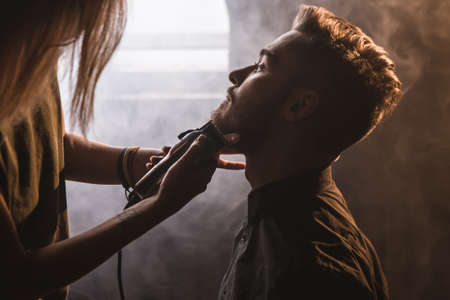 Female hairdresser shaving stylish man closeup. Atmospheric barbershop with white smoke, young fashionable male in focus on foreground, unrecognizable woman with electric razorの写真素材