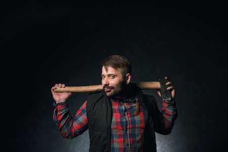 Strong smirking seductive worker holding axe. Adult woodsman on dark background, confident lumberjack portrait closeup. Sexy rural male, strength conceptの写真素材