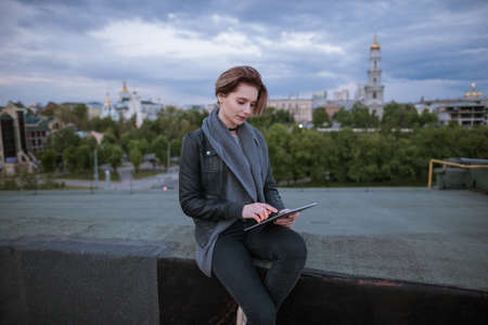 Girl with tablet working on city background. Smart woman sitting on the buildings roof in city during work. Blogger, distant work, wireless internet conceptの写真素材