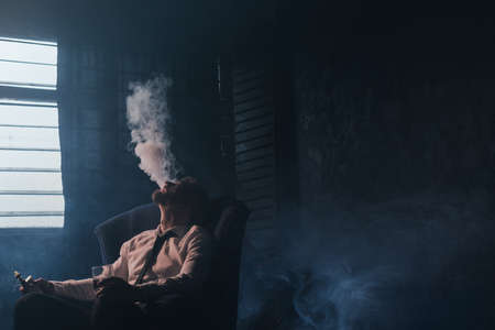 Businessman relaxing with vape and alcohol. Dark room with white atmospheric smoke background, rich people entertainment, private club for luxury life with free spaceの写真素材