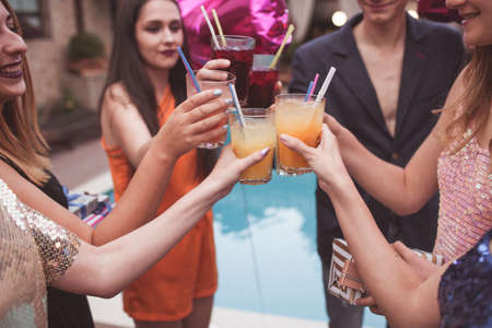 Celebration party. Joyful friends company with alcohol cocktails. Modern youth with drinks in focus on foreground closeup. Happy birthday toast, tasty beverages, congratulation conceptの写真素材