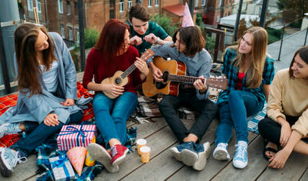 Group of friends listen to buskers singing and playing guitar and ukulele at a roof top party. Musician artistic lifestyle. Teenagers leisureの写真素材