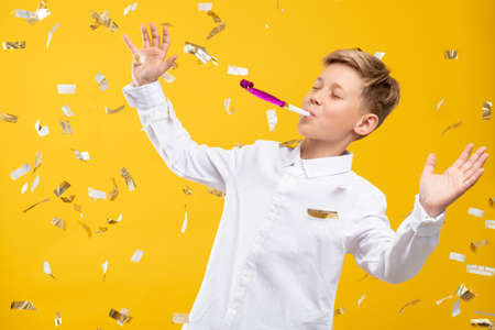 Birthday boy portrait. Fun celebration. Amused kid blowing party horn in confetti rain isolated on orange background.の写真素材