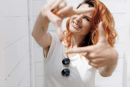 Possitive redhead woman in sunglasses doing frame outdoors on white backgroundの写真素材