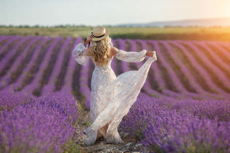 pretty blonde woman running away in lavender field. woman in long dress and straw hat having fun in flowers of lavenderの写真素材