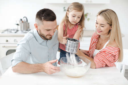 Cute little girl and her beautiful parents are cooking cake at kitchen