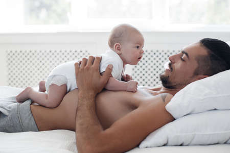 man with infant. happy family at home. happy father with cute two months baby boy laying in light bed at homeの写真素材
