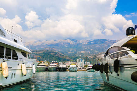 Luxury yachts and sailing ships moored at wharf in Budva marina, Montenegro. Port in Mediterranean sea. White motor boats and sailboats docked in harbor against mountains. Fashionable vacation.の写真素材