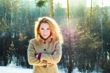 Smiling girl in the winter wood at sunset. Beautiful young woman wearing coat with curly blonde hair posing in a snowy forest at sunny evening. Caucasian female model looking at camera. Seasonal fashion.の写真素材