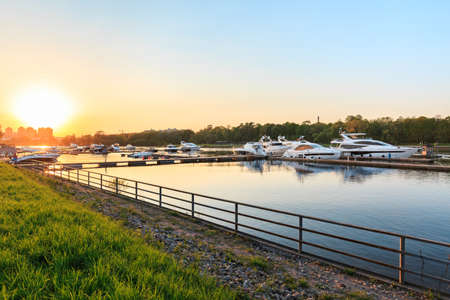 Luxury yachts docked in marina at colorful sunset. Marine parking, modern motor boats moored in water at pier. Summer view of Neva river bank, embankment and superyachts in sunshine. Sunny evening in St. Petersburg, Russia. Travel and fashionable vacation.の写真素材
