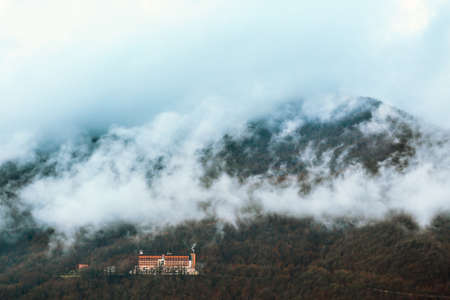 Forested mountain slope shrouded in mist. Foggy landscape view in autumn. Fog enveloping forest with bare winter trees. Lonely hotel in woods. Mystical place. Silence.の写真素材