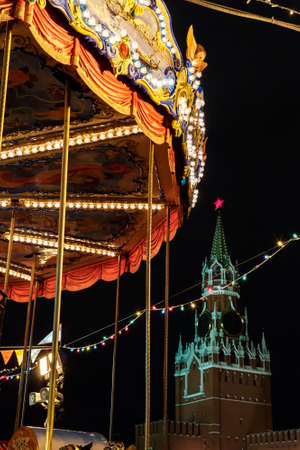 Merry Go Round and Moscow Kremlin illuminated at night by Christmas lights. Vintage colorful carousel flying horse at christmas funfair on red square outdoors in winter holidays. Russia.の写真素材