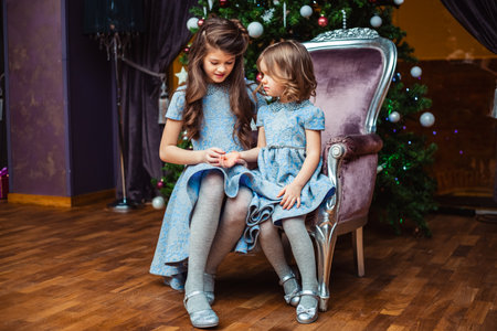 St. Petersburg, Russia - January 22, 2017: Portrait of two sisters wearing beautiful dresses sitting in chair in living room with traditional Christmas tree decorations to winter holidays.のeditorial素材