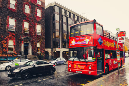Dublin, Ireland - November 6, 2018: Red double-decker city tour bus waiting for tourists at stop on rainy day. Brown vintage block walls, historic place, walking people. Romantic scene in fall season.のeditorial素材