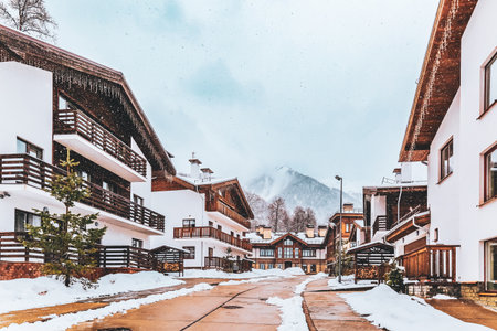 Cozy wooden cottages in chalet style covered with snow. Beautiful snowcapped mountain peak. Xmas picturesque background. Rosa Khutor, Krasnaya Polyana, Sochi, Russia.の写真素材