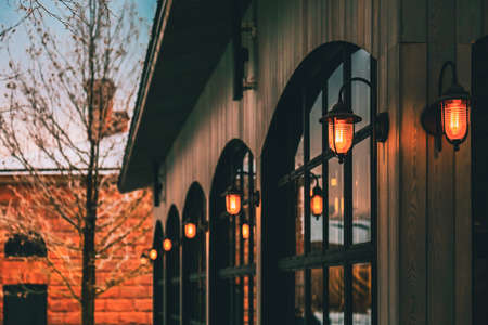 One-story building. Row of black arched windows in dark wooden wall. Vintage lanterns close-up. Orange stone house and leafless bare tree branches covered by snow at background.の写真素材