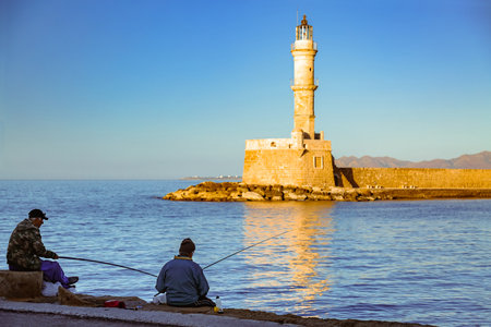 Chania, Crete, Greece â November 3, 2021: Two fishermen on embankment. The Venetian Lighthouse in old harbor at sunset. Mediterranean Seaのeditorial素材