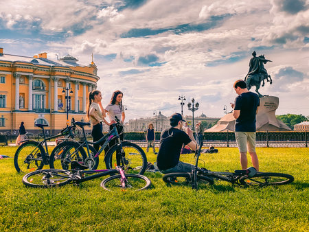 St. Petersburg, Russia - June 7, 2020: Sporty male and female friends rest after cycling in sunny day. Bikes parked on grass. Statue of Peter Great and Constitutional Court at backのeditorial素材