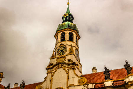 Exterior facade of Loreta church in Prague: green rooftop on white belfry, white walls, red rooftop. In a cloudy gloomy day, green trees parkの写真素材