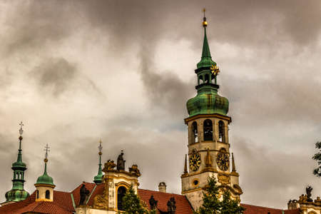 Exterior facade of Loreta church in Prague: green rooftop on white belfry, white walls, red rooftop. In a cloudy gloomy day, green trees parkの写真素材