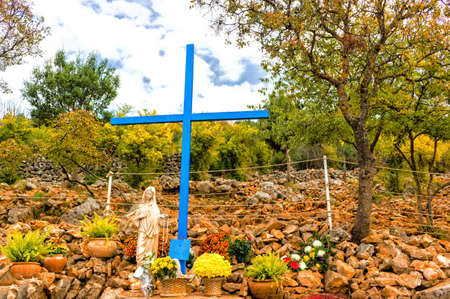 The Blue Cross and Statue of Blessed Virgin Mary at Apparition Mountain in Medjugorje among grey rocks and green trees and weedsの写真素材