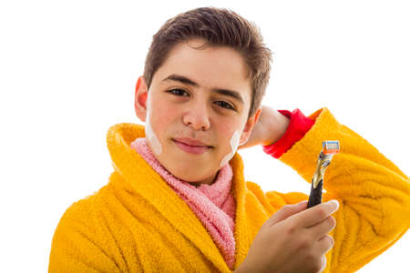 A Hispanic boy wears a yellow bathrobe with a pink towel around his neck: he has some patches on his face but smiles holding the razor he used for shavingの写真素材