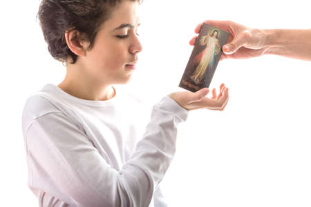 A happy Caucasian smooth-skinned boy in a white long sleeves t-shirt smiles receiving with right hand a Merciful Jesus wood icon given by an adult handの写真素材