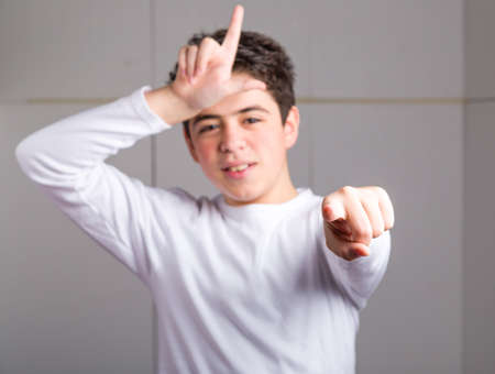 Blurred Hispanic teen with acne skin in a white long sleeve t-shirt smiles making loser gesture with...