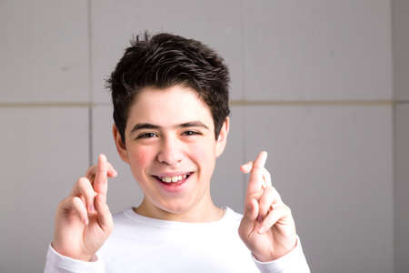 Latin young Smooth-skinned boy in a white long sleeve t-shirt smiles crossing fingers of both hands as superstitious  gesture to get luck on an industrial backgroundの写真素材