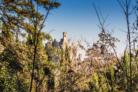 A  medieval crenellated clock tower stands on a peak overlooking the valley of a village and a country of farmland, bushes, cypresses and other tress among braches in the mist of a sunny winter dayのeditorial素材